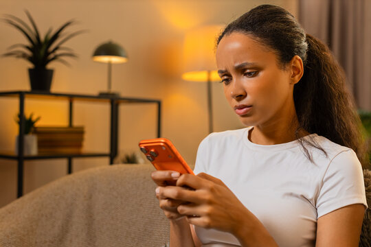 African American young woman on home sofa checks suspicious message then deletes it after spotting fraud. Black girl blocks sender and protects account during home smartphone browsing safely indoors.
