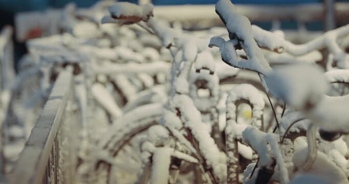 A row of bicycles covered in snow on public parking at night. The snow is piled up on the handlebars and the seat of the bikes. The bikes are leaning against a fence