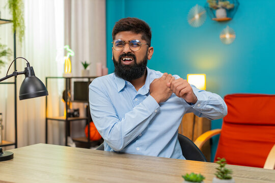 Indian man at home clutches shoulder wincing from sharp severe pain possible injury strained muscle discomfort. Arabian guy at desk grimaces seeking massage and rest hopes for recovery pain ease soon.