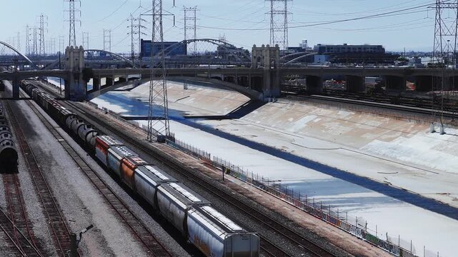 Aerial shot follows a long freight train moving along railroad tracks adjacent to the concrete Los Angeles River channel near the historic Fourth Street Bridge under clear sky.