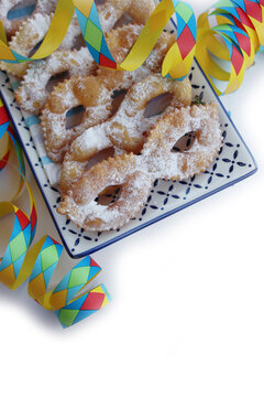 Italian sweet crisp pastry deep-fried and sprinkled with sugar in shape of Carnival mask  in a ceramic tray isolated on white background. Chiacchere or crostoli, bugie, cenci with colorful paper serpe