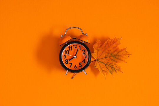 Autumn conceptual background with orange alarm clock showing midnight and dry maple leaf on vibrant orange surface. Flat lay seasonal fall layout or daylight saving time concept with copy space.