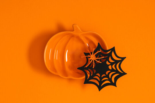 Halloween composition with pumpkin shaped ceramic plate, decorative black spider web and small orange spider on bright background. Flat lay modern festive layout with blank copy space for text.