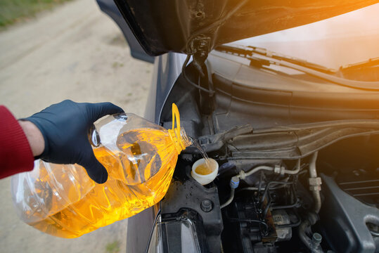 Gloved hand pours bright yellow winter washer fluid from large plastic bottle into windscreen reservoir of car, preparing vehicle for winter driving. Filling car washer fluid container from large jug