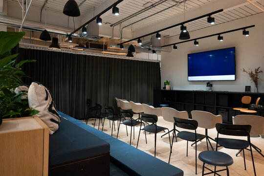 View of a modern office meeting room featuring rows of chairs, a large wall-mounted screen, dark curtains, and indoor plants in London, United Kingdom.