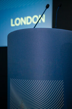 View of a blue podium with a modern white line pattern and two microphones, set before a screen displaying the word London in London, United Kingdom.