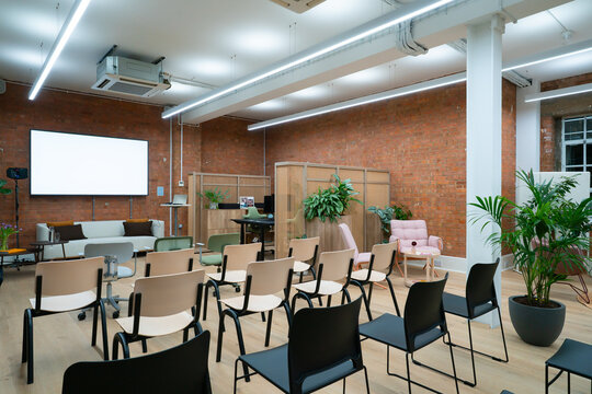 View of a modern office meeting room with rows of chairs, a white projection screen, exposed brick walls, and indoor plants in London, United Kingdom.