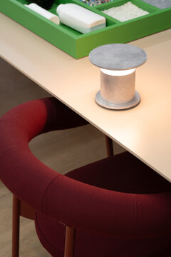 View of a modern cylindrical desk lamp with a glowing light on a clean table surface next to a green tray, with a red upholstered chair in the foreground in London, United Kingdom.