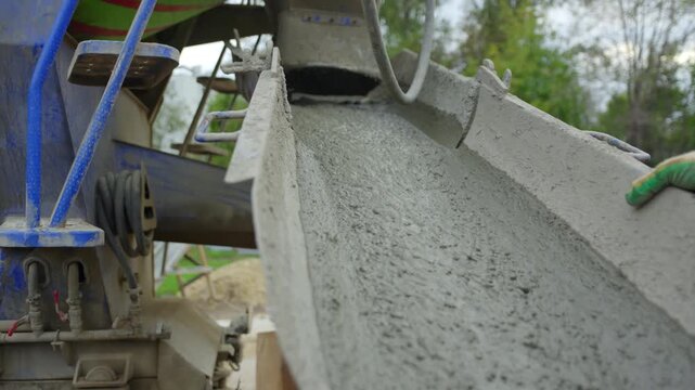 Long metal chute of mixer truck delivers fresh concrete mix into frame. Close perspective displays steady flow of industrial cement slurry