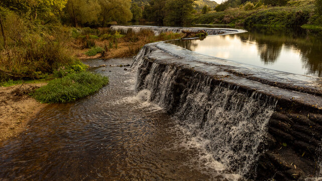 Water flows over the Warleigh Weir at sunset in a lush green valley.