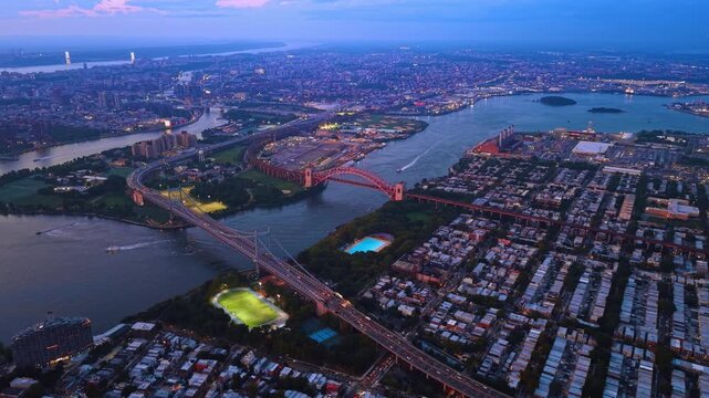 New York bridges connect riverbanks beside glowing sports fields. A red arch and suspension towers rise above blue water with rows of houses in the foreground.