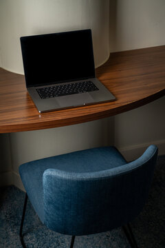 View of a modern laptop placed on a curved wooden desk with a blue upholstered chair in a contemporary workspace in London, United Kingdom.