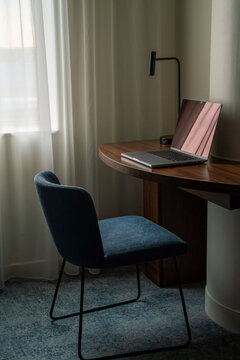 View of a modern laptop placed on a curved wooden desk with a blue upholstered chair and a minimalist desk lamp in a bright room with sheer curtains in London, United Kingdom.