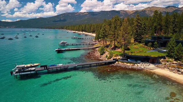 Long piers extend from a wooded beach into Lake Tahoe. Boats rest beside docks in turquoise water below pine trees and distant mountains under white clouds.
