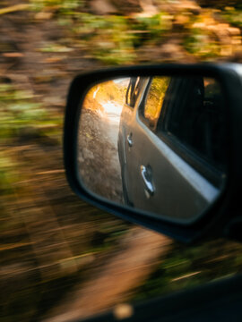 View of the side mirror of a moving vehicle reflecting a sunlit road and landscape during golden hour in Kecamatan Kambera, Nusa Tenggara Timur, Indonesia.