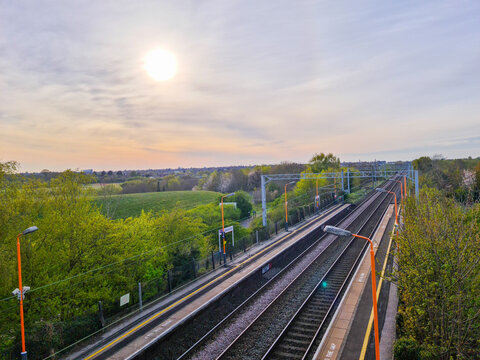View of Marston Green Station with railway tracks, platforms, and overhead lines surrounded by lush green trees under a sunset sky in Marston Green, England, United Kingdom.