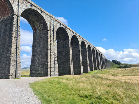 View of the historic Ribblehead Viaduct with its massive stone arches stretching across the grassy landscape under a bright blue sky in Carnforth, England, United Kingdom.