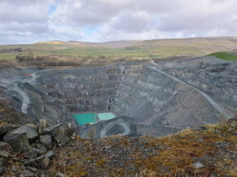 View of a large open-pit quarry with terraced rock walls and turquoise water pools, set against the rolling hills of the Yorkshire Dales National Park, England, United Kingdom.