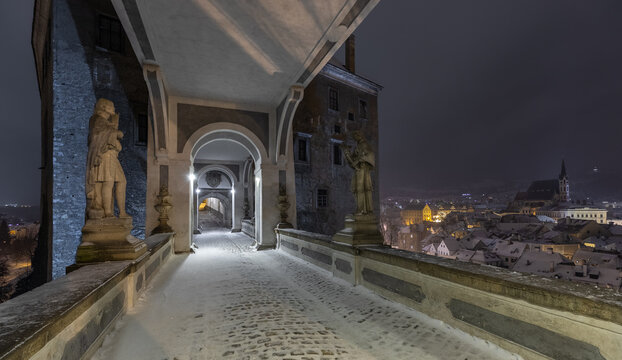 View of the snow-covered Cloak Bridge at Cesky Krumlov Castle with historic statues and illuminated arches overlooking the town rooftops at night in Cesky Krumlov, South Bohemian Region, Czechia.