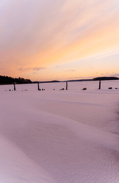View of a frozen, snow-covered bay with wooden poles protruding from the ice under a soft pastel sunset sky in Raasepori, Uusimaa, Finland.
