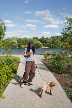 A person walks a dog while checking their phone near a lake