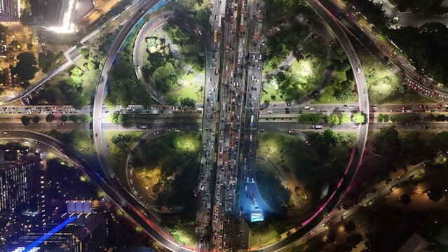 Aerial view of the Semanggi Interchange at night, featuring illuminated cloverleaf ramps, heavy traffic flow, and surrounding urban greenery in Jakarta Selatan, DKI Jakarta, Indonesia.