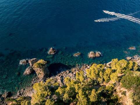Aerial view of the rugged coastline with turquoise waters, rocky outcrops, and lush vegetation, featuring two motorboats leaving white wakes on the sea, Positano, Campania, Italy.