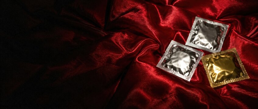 Silver and gold condom packets on red satin fabric with dark moody lighting and copyspace