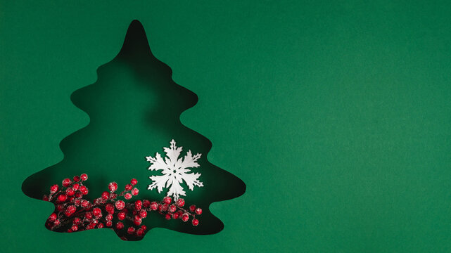 Minimalist Christmas background with a tree cutout in green paper, decorative white snowflake, and red berries. Festive winter composition with copy space for New Year greeting card text.