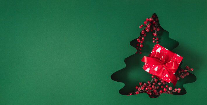 Minimalist Christmas banner with a tree cutout in green paper, red gift box with ribbon bow, and frosted berries. Festive winter background with large copy space for New Year greeting text.