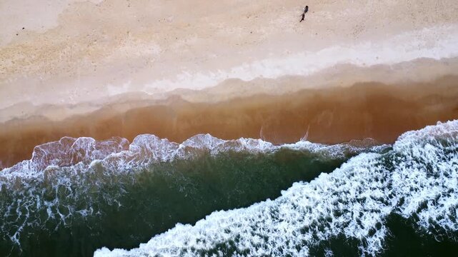 Aerial drone footage moving upward over a sandy beach as gentle ocean waves roll onto the shore, creating intricate white foam patterns and beautiful natural textures along the coastline.