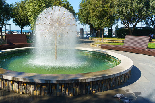A circular stone fountain with a distinctive radial water spray stands in Hlavatyjev Park in Koper, Slovenia