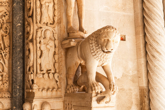 Intricately carved medieval stone sculpture at the entrance of Trogir Cathedral, depicting a standing female figure above a lamb alongside richly ornamented Gothic reliefs and twisted columns