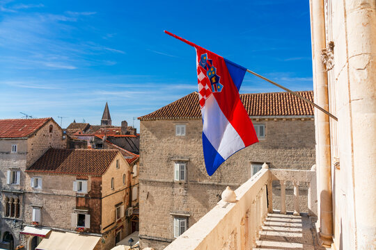 Croatian flag waving in the foreground above the historic center of Trogir, with the town&rsquo;s medieval stone architecture, red-tiled roofs and distinctive clock tower visible against a bright blue sky