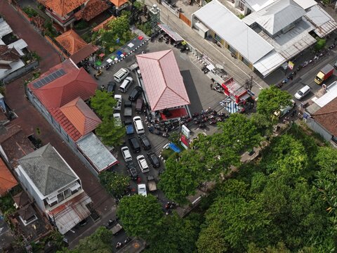 Aerial view of SPBU 54.803.11 Abianbase gas station with cars and motorcycles queuing for fuel, surrounded by lush trees and residential buildings in Kecamatan Mengwi, Bali, Indonesia.