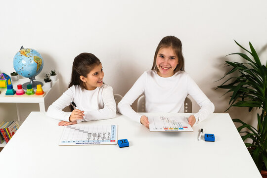 Two smiling schoolgirls sitting at a table and practicing mathematics with visual learning boards for multiplication and division in a bright classroom.