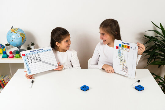 Two schoolgirls smiling at each other while holding educational boards with multiplication and division exercises during a mathematics lesson.