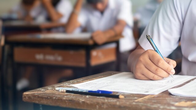 Student Writing Exam Paper with Stressed Classmates in Background Classroom