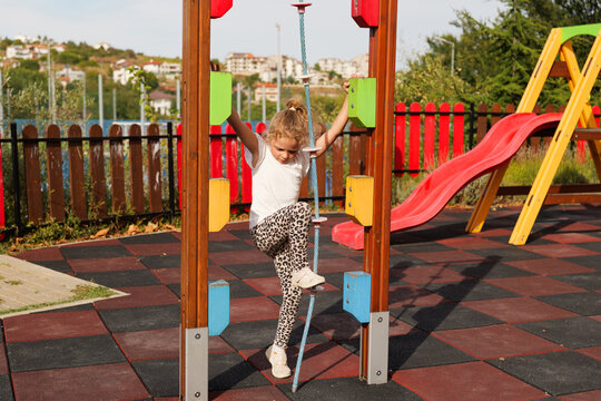 Little girl climbing up a climbing rope on an outdoor playground with a colorful slide in the background.