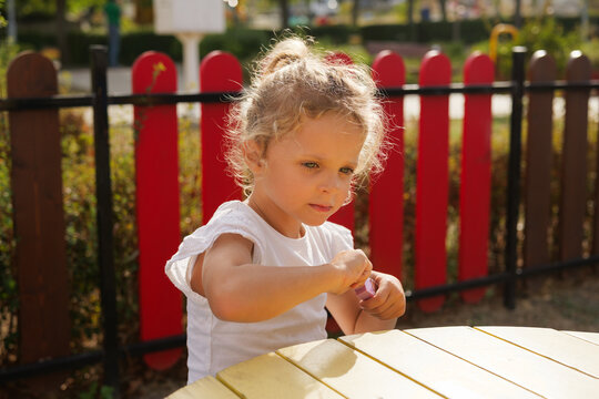 Little girl with blonde curly hair sitting at a wooden table outdoors and playing with cosmetics.