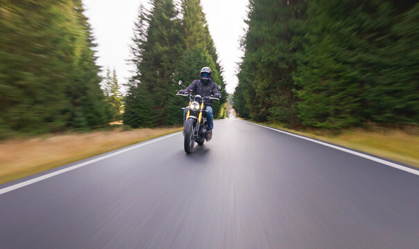 A rider on a stylish retro motorcycle travels along a paved forest road in the evening. Shot in motion with a panning camera, the blurred asphalt and soft dusk light create a dynamic sense of speed.
