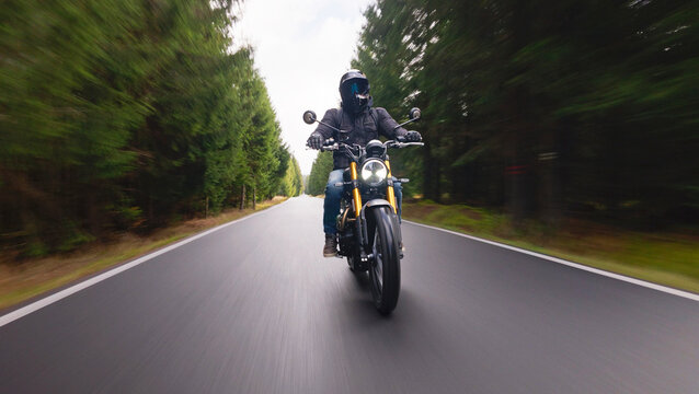 A rider on a stylish retro motorcycle travels along a paved forest road in the evening. Shot in motion with a panning camera, the blurred asphalt and soft dusk light create a dynamic sense of speed.