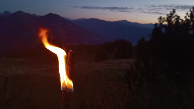 A burning wooden torch glowing brightly against a dark alpine mountain landscape at dusk, capturing a traditional ritual fire event under a serene evening sky in the mountains.