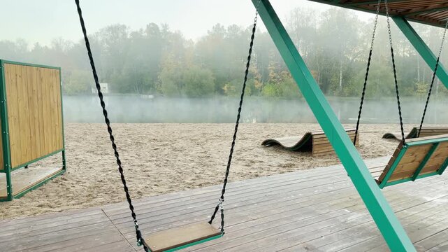 Tracking shot of an empty lakeside beach park on gimbal. Smooth sideways camera movement passes wooden swings near a foggy lake in Pushkin, Russia. The quiet scene evokes calm nostalgia in slow motion