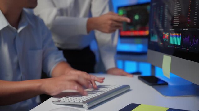 Close up of traders hands typing on keyboard and colleagues discussing stock market charts on a computer in a modern office at night