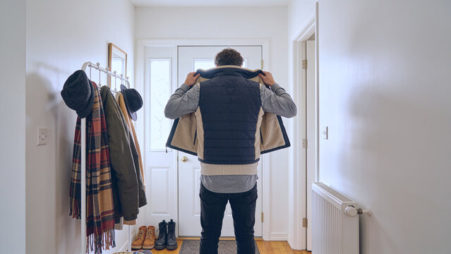 A man puts on a warm fleece-lined quilted vest in a bright home entryway, standing near a coat rack filled with jackets