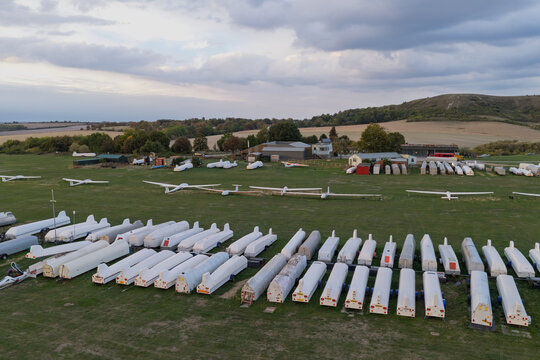 Aerial view of a glider airfield featuring numerous gliders parked on the grass and rows of white trailers, set against a backdrop of rolling hills and a cloudy sky in the United Kingdom.