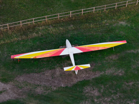 Aerial view of a glider with red, yellow, and white wings parked on a grassy field next to a wooden fence, United Kingdom.