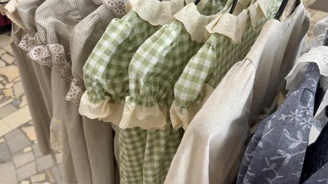 Vintage style fabric garments displayed on rack, featuring green gingham, cream linen, and blue floral embroidery with lace trim, useful for boutique fashion, textile, and clothing retail