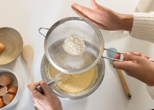 Diverse female friends sifting flour into yellow batter on white countertop using fine-mesh sieve